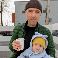 White man in winter hat and green jacket holding a coffee cup and a baby with pacifier.